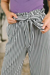 A close-up of a person wearing black and white vertically striped high-waisted pants with a matching fabric tie belt, paired with a purple ribbed top.