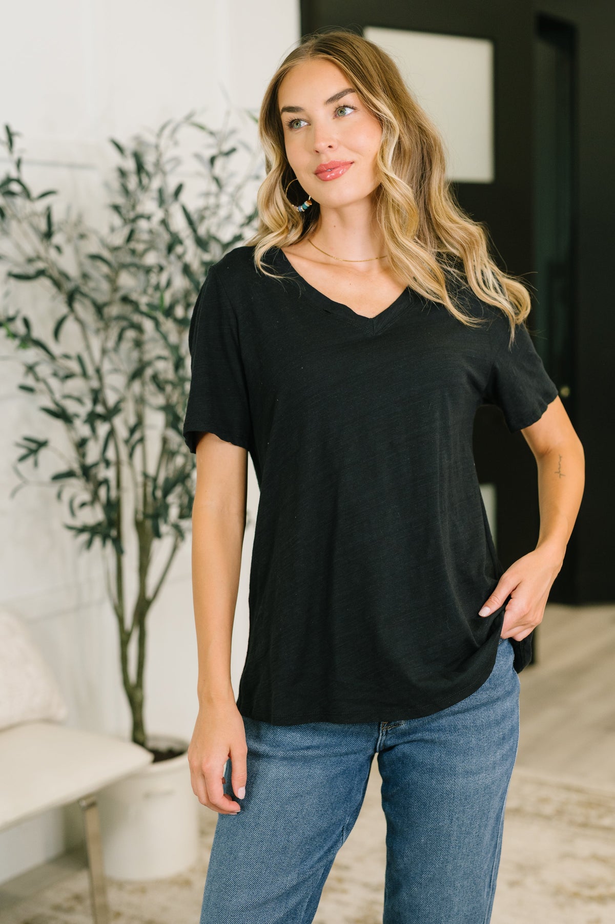 Woman wearing a black t-shirt and blue jeans standing indoors with a plant in the background