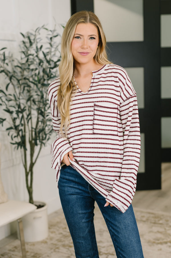 A woman modeling a long-sleeve knit top with horizontal burgundy and off-white stripes, paired with blue jeans, in a brightly lit indoor setting.