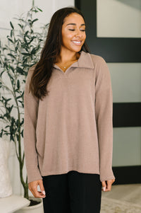 Woman wearing a beige ribbed sweater standing indoors with a plant in the background