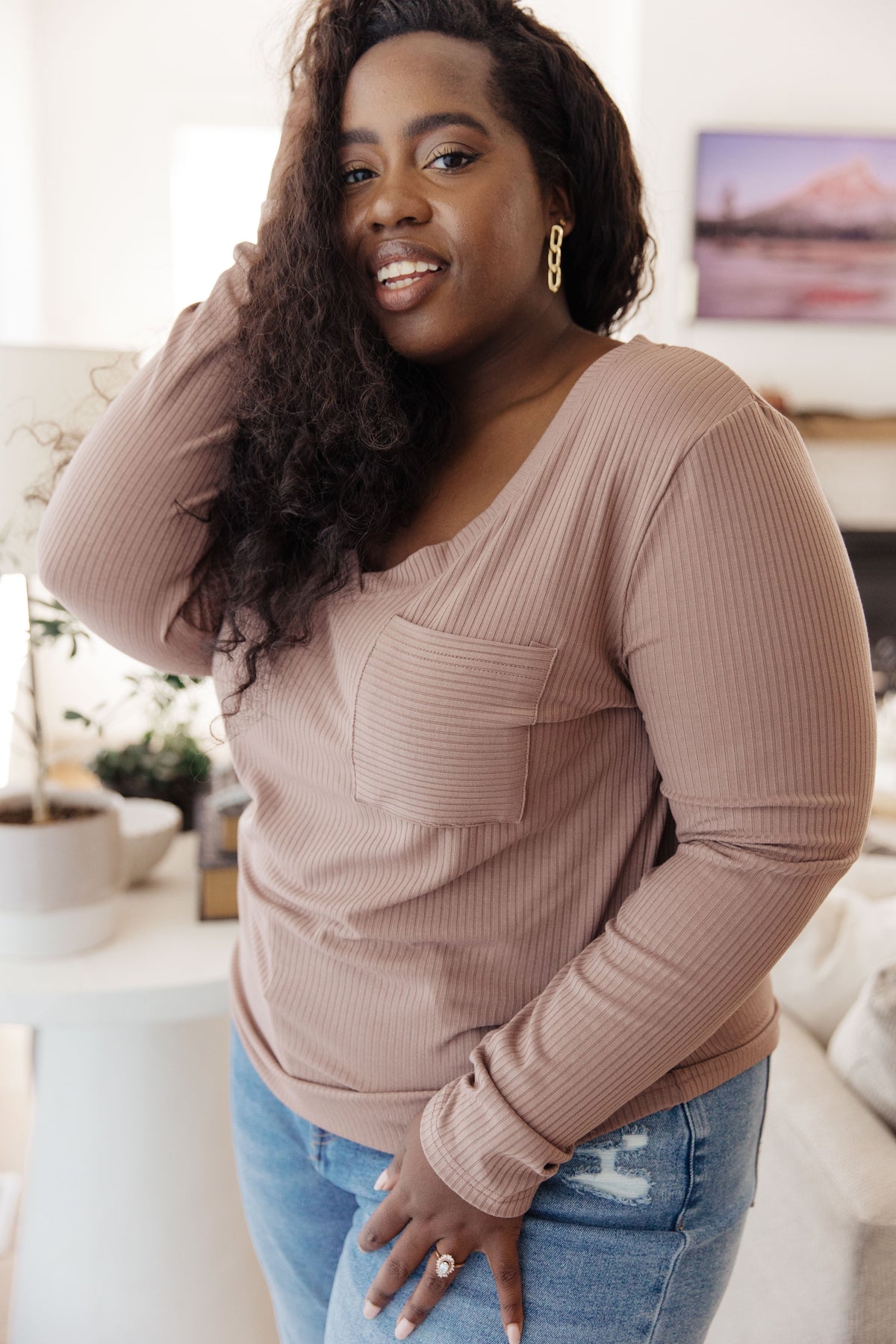A woman wearing a ribbed long-sleeve scoop-neck top in a dusty rose color, paired with distressed blue jeans, standing in a cozy living room setting.