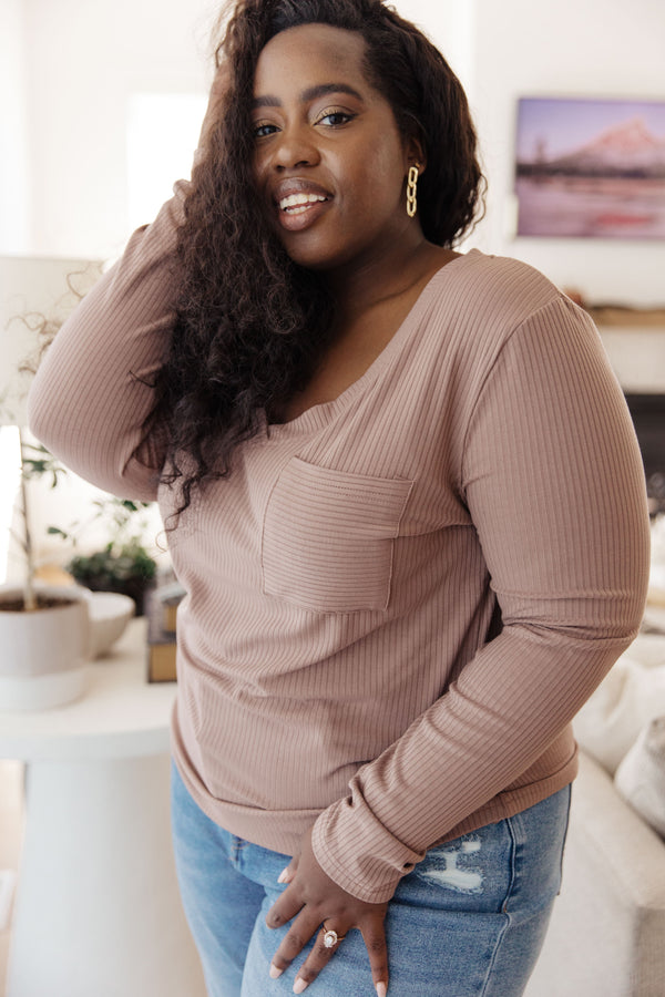 A woman wearing a ribbed long-sleeve scoop-neck top in a dusty rose color, paired with distressed blue jeans, standing in a cozy living room setting.