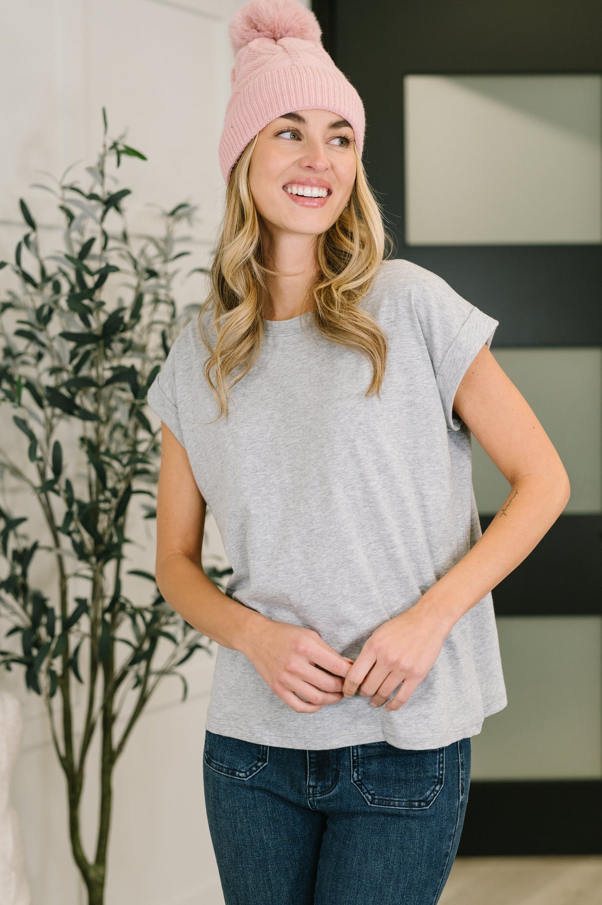 Woman wearing a gray t-shirt, pink beanie, and blue jeans standing indoors.