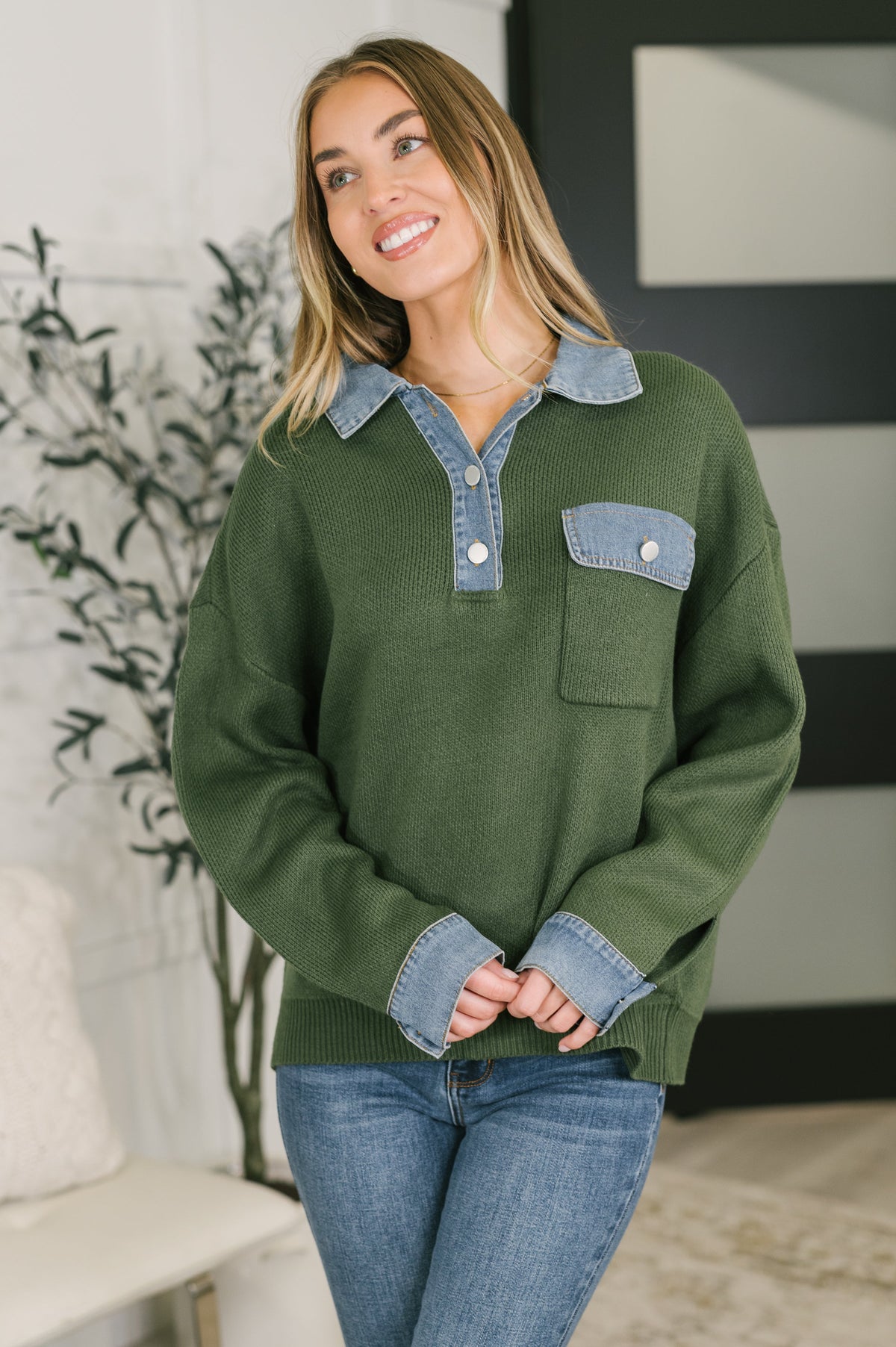 A woman wearing an olive green knit sweater with a light blue denim collar and chest pocket, paired with blue jeans, standing in a modern indoor setting.