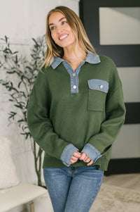 A woman wearing an olive green knit sweater with a light blue denim collar and chest pocket, paired with blue jeans, standing in a modern indoor setting.
