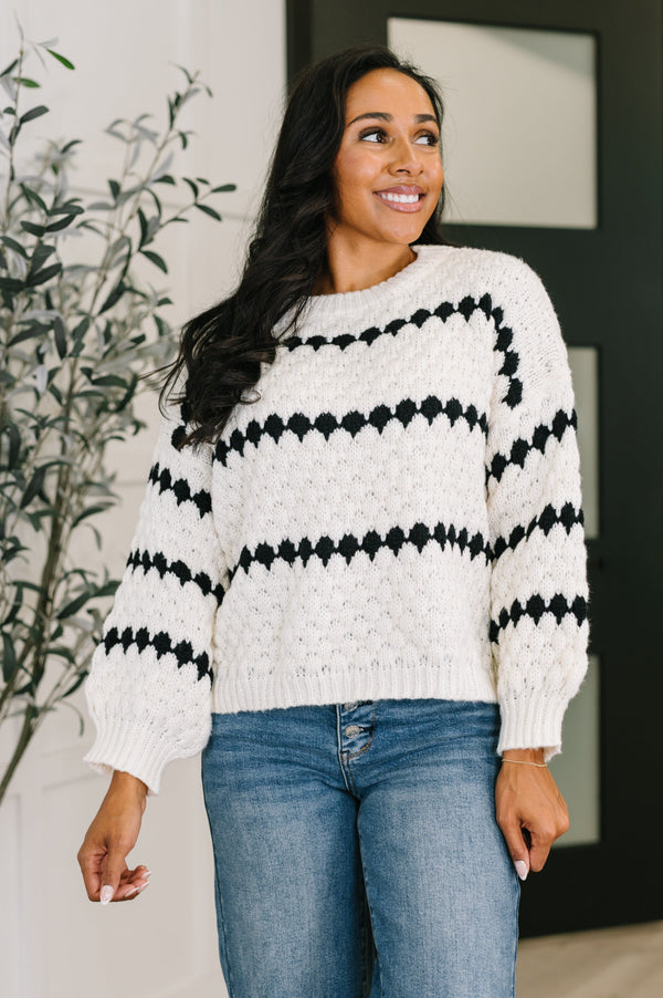 Woman wearing a white sweater with black pattern and blue jeans indoors.