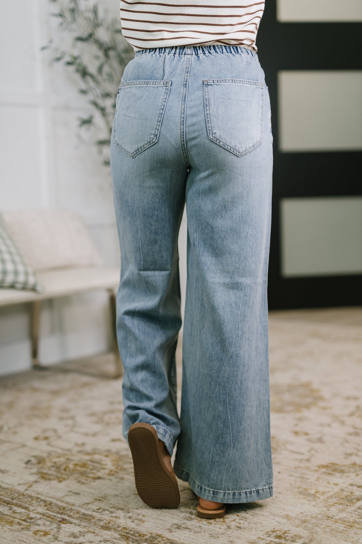 Person wearing light blue jeans standing in a room with a plant and bench in the background.