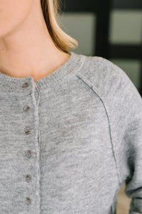Close-up of a person wearing a light heather gray knit cardigan with a buttoned front and raglan sleeves.