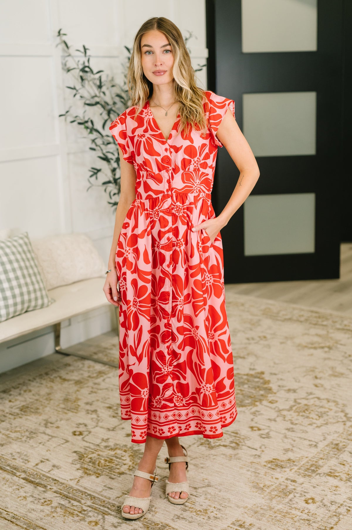 Woman wearing a red floral dress standing in a room with a couch and plant.