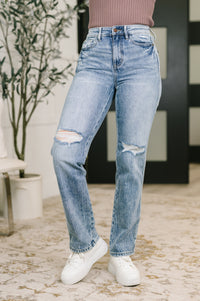 Person wearing light blue ripped jeans and white sneakers in a room with a plant and decorative items.