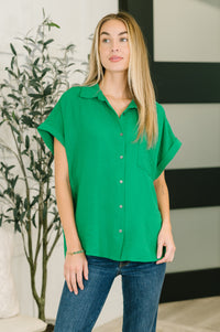 Woman wearing a green shirt and blue jeans standing indoors with a plant in the background