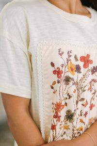White t-shirt with a floral design on the chest pocket against a neutral background