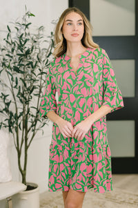 Woman wearing a pink and green patterned dress standing indoors with a plant in the background.