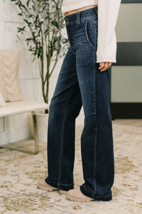 A person wearing dark blue wide-leg jeans, a white long-sleeve top, and beige boots, standing on a patterned rug in a modern indoor setting.