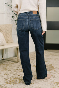 A person shown from the back wearing dark blue flare jeans with a cream sweater, standing on a patterned beige rug in an indoor room setting.