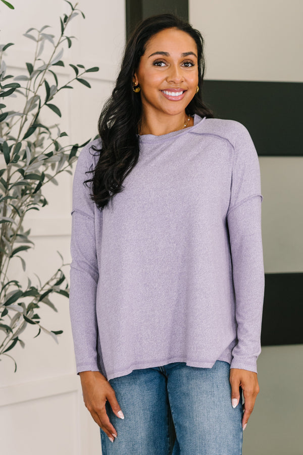 Woman wearing a light purple sweater and blue jeans standing indoors with a plant in the background