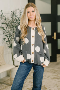 A woman wearing a gray cardigan with large white polka dots and cream ribbed trim, paired with blue jeans, standing in a modern indoor setting.