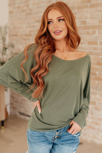 Woman wearing an olive green long-sleeve top and blue jeans against a brick wall.
