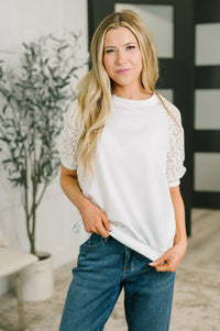 A woman wearing a white short-sleeve top with textured lace sleeves and blue jeans, standing in a brightly lit indoor setting.
