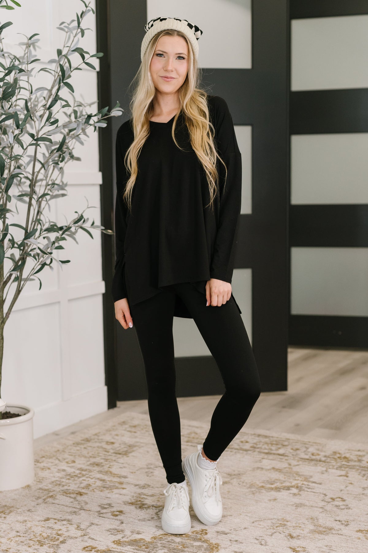 Woman wearing a black outfit with white sneakers in a room with a plant and striped wall.