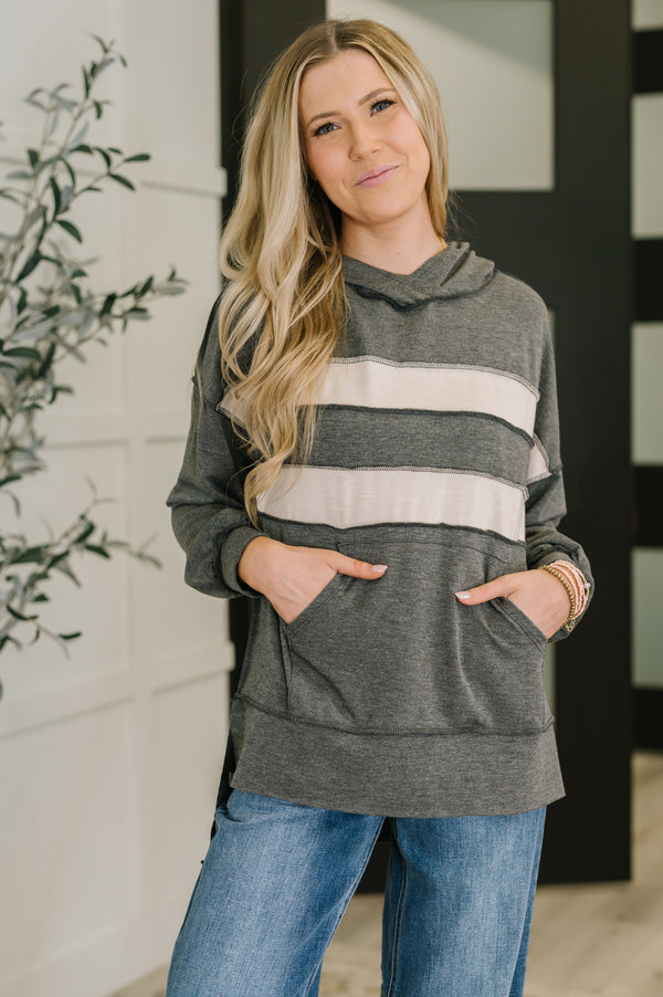 Woman wearing a gray hoodie with white stripes and blue jeans, standing indoors.