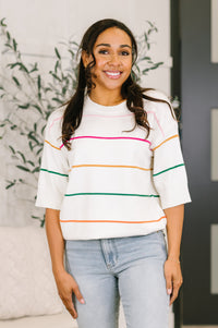 Woman wearing a white sweater with colorful stripes and light blue jeans indoors.