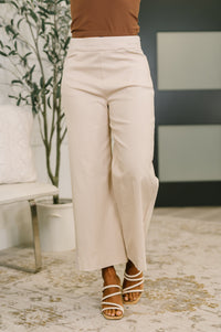 Person wearing beige wide-leg pants and brown top in a room with a plant and chair.