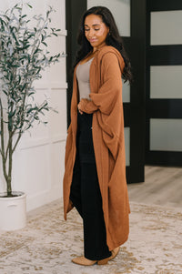 Woman wearing a long brown cardigan over a white top and black pants, standing indoors.