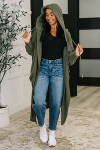 Woman wearing a long green cardigan, black top, and blue jeans indoors.