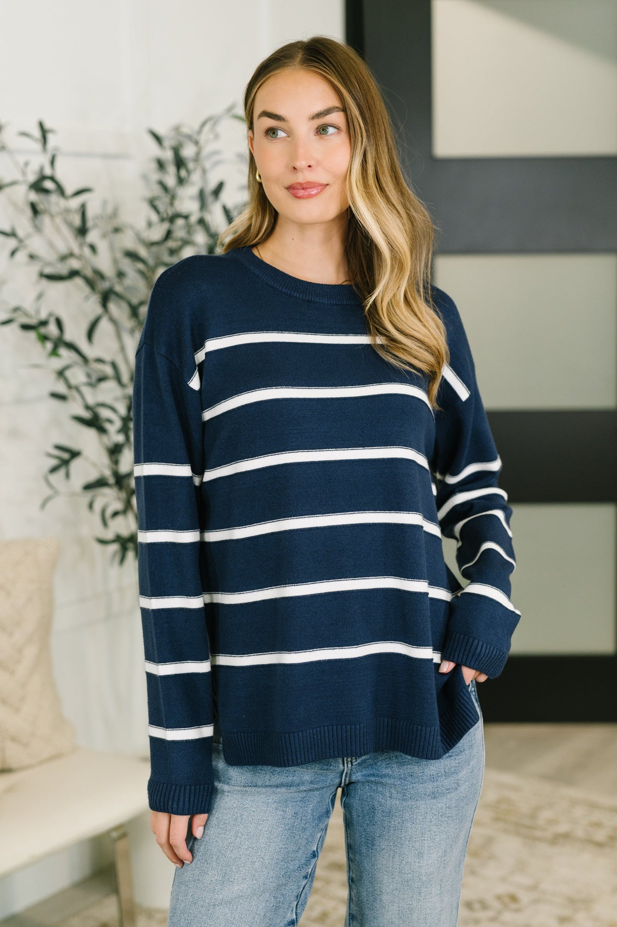 A woman wearing a navy blue and white horizontally striped knit sweater with light blue jeans, standing in a brightly lit indoor setting.