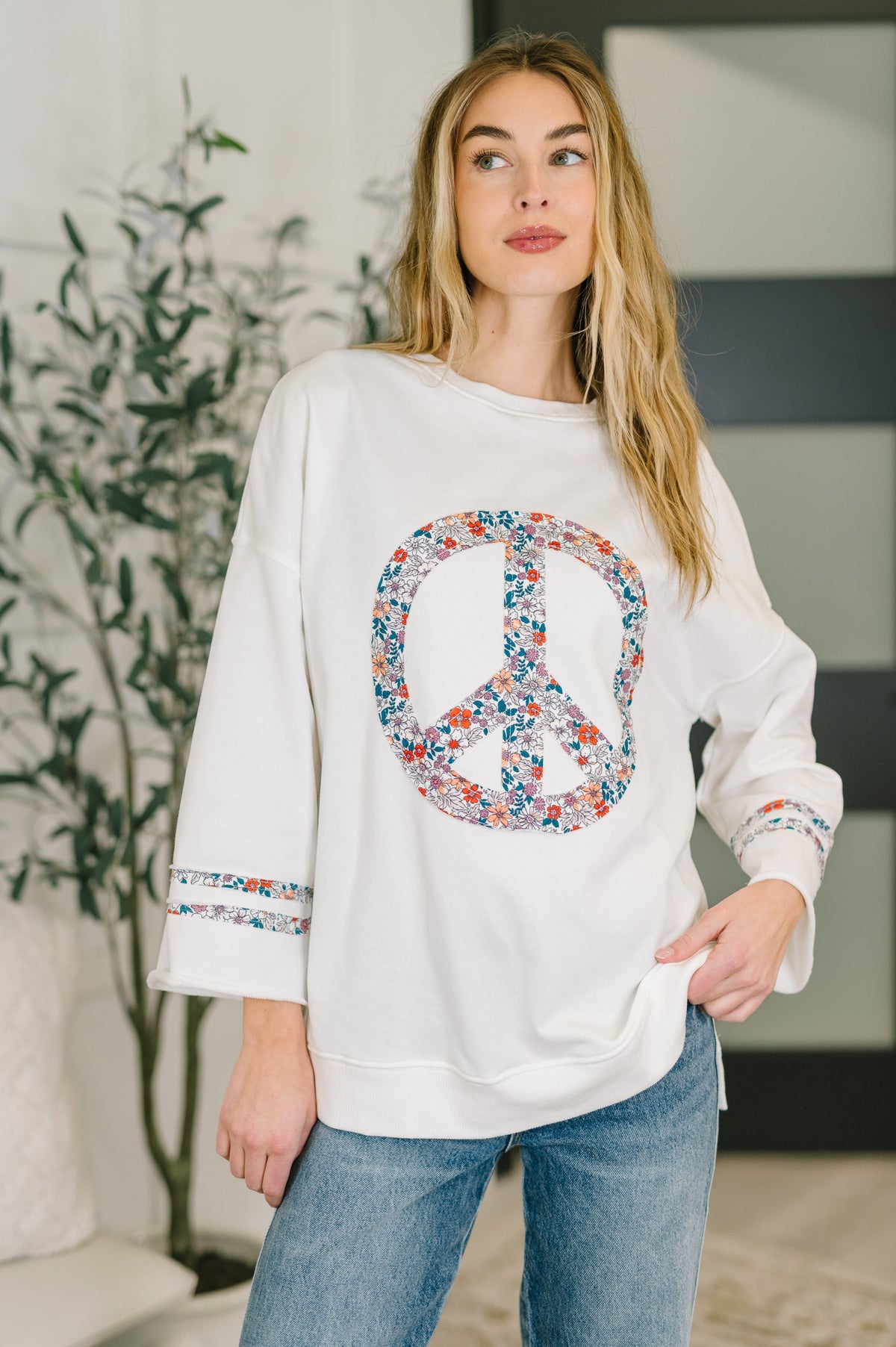 Woman wearing a white sweatshirt with a colorful peace symbol design, standing indoors.