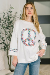 Woman wearing a white sweatshirt with a colorful peace symbol design, standing indoors.
