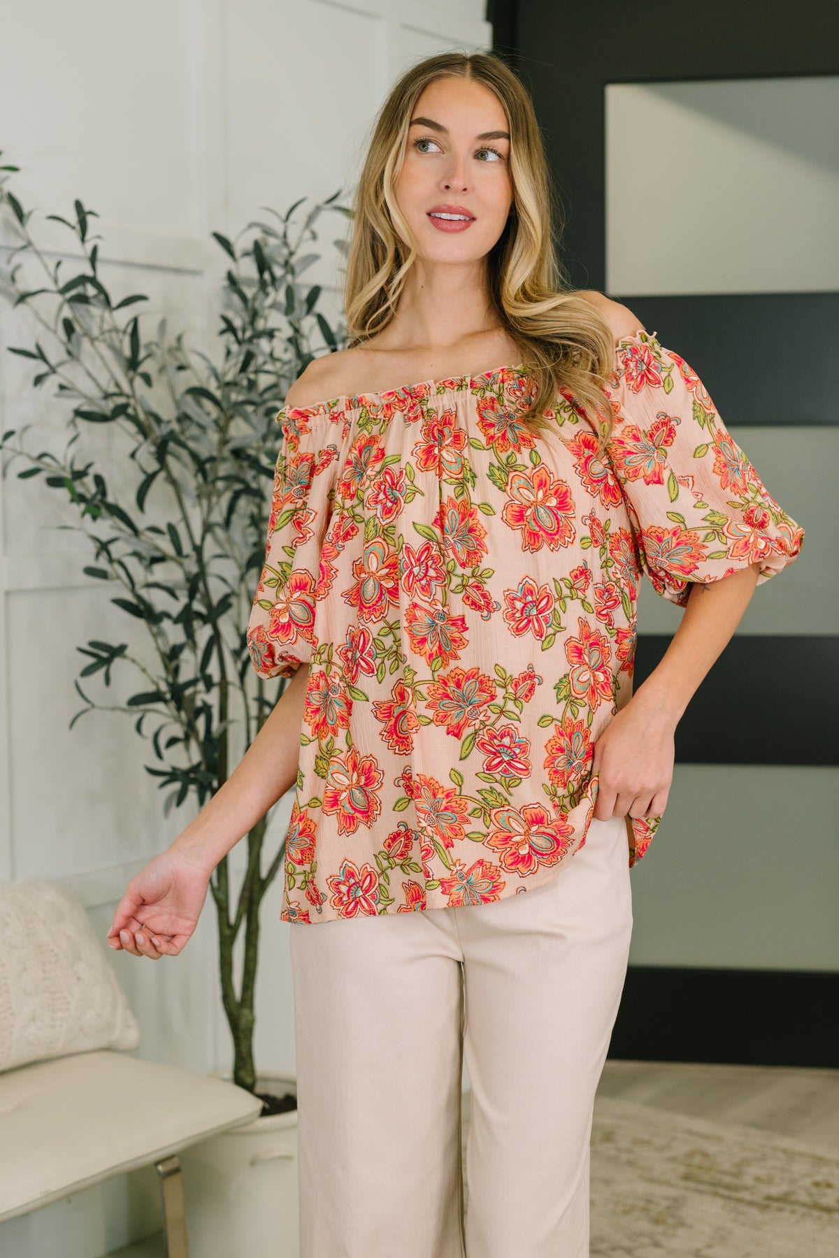Woman wearing a floral off-shoulder top in a room with a plant and couch.
