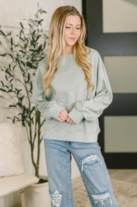 Woman wearing a light gray sweatshirt and blue jeans in a living room setting.