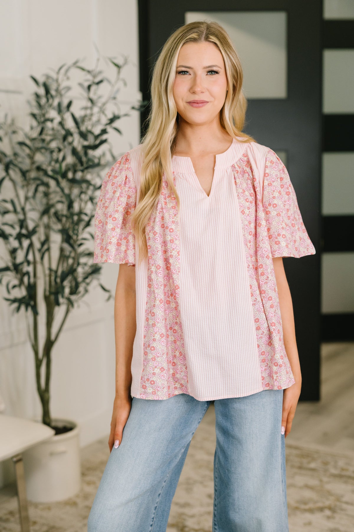 A woman modeling a light pink short-sleeve top with a vertical textured center panel and pink floral ruffle sleeves, paired with light blue jeans in an indoor setting.