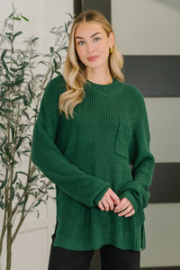 Woman wearing a green sweater standing indoors with a plant in the background