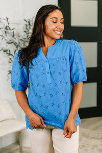 Woman wearing a blue blouse with floral embroidery in a room with a plant and white wall.