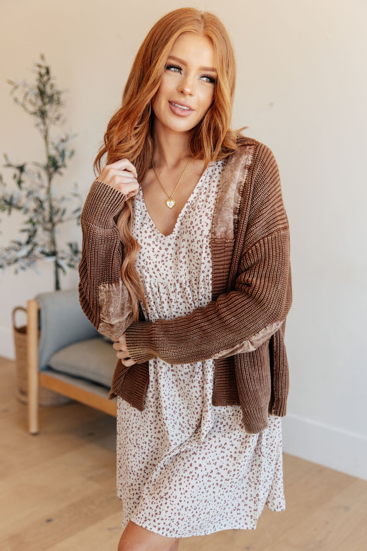 A woman with long red hair stands indoors wearing a brown open-front knit cardigan over a white V-neck dress with a small brown allover print. The photo is taken in a softly lit room with a light wall, wood floor, and a chair in the background.