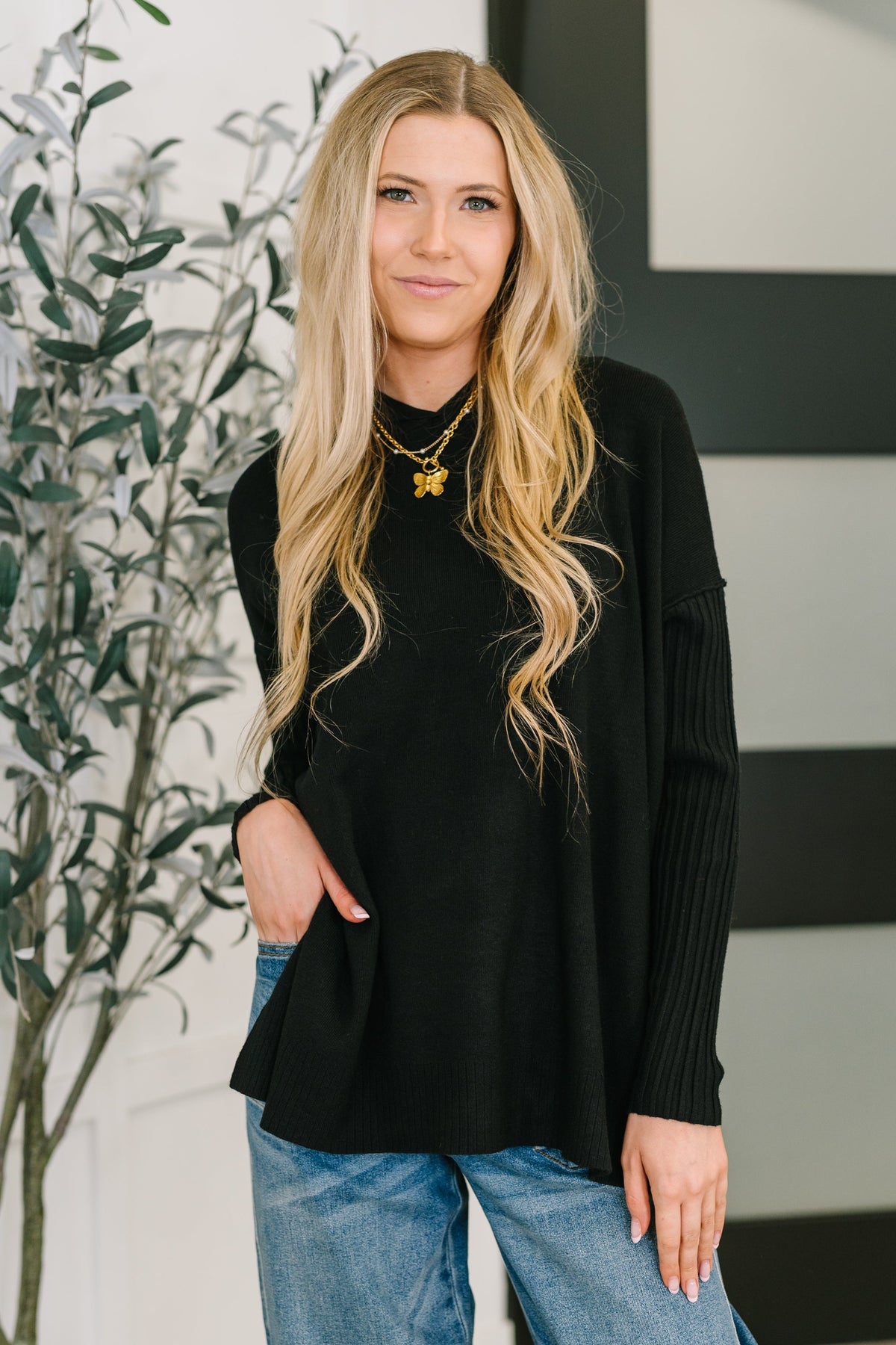 Woman wearing a black sweater and blue jeans standing indoors with a plant in the background