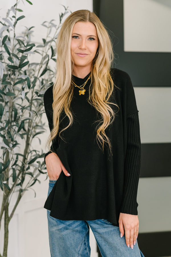 Woman wearing a black sweater and blue jeans standing indoors with a plant in the background