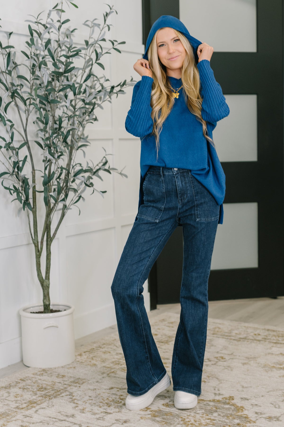 Person wearing a blue hoodie and jeans standing indoors with a plant in the background