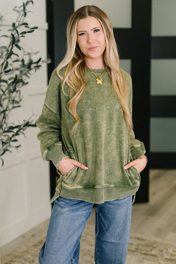 Woman wearing a green sweatshirt and blue jeans standing indoors with a plant in the background