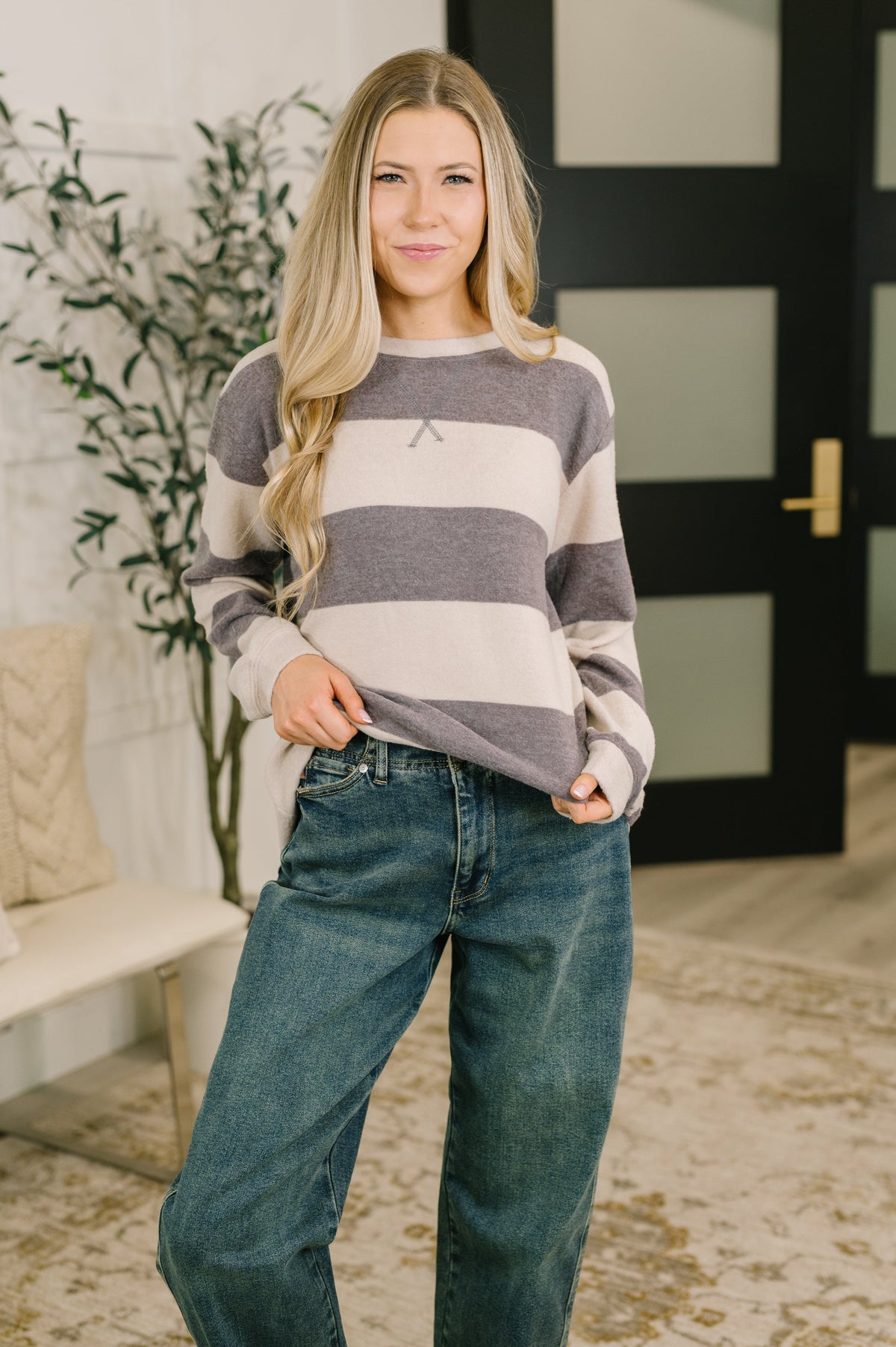 A woman wearing a gray and cream striped long-sleeve sweater with a small embroidered logo on the chest, paired with blue jeans, standing in a modern indoor setting.