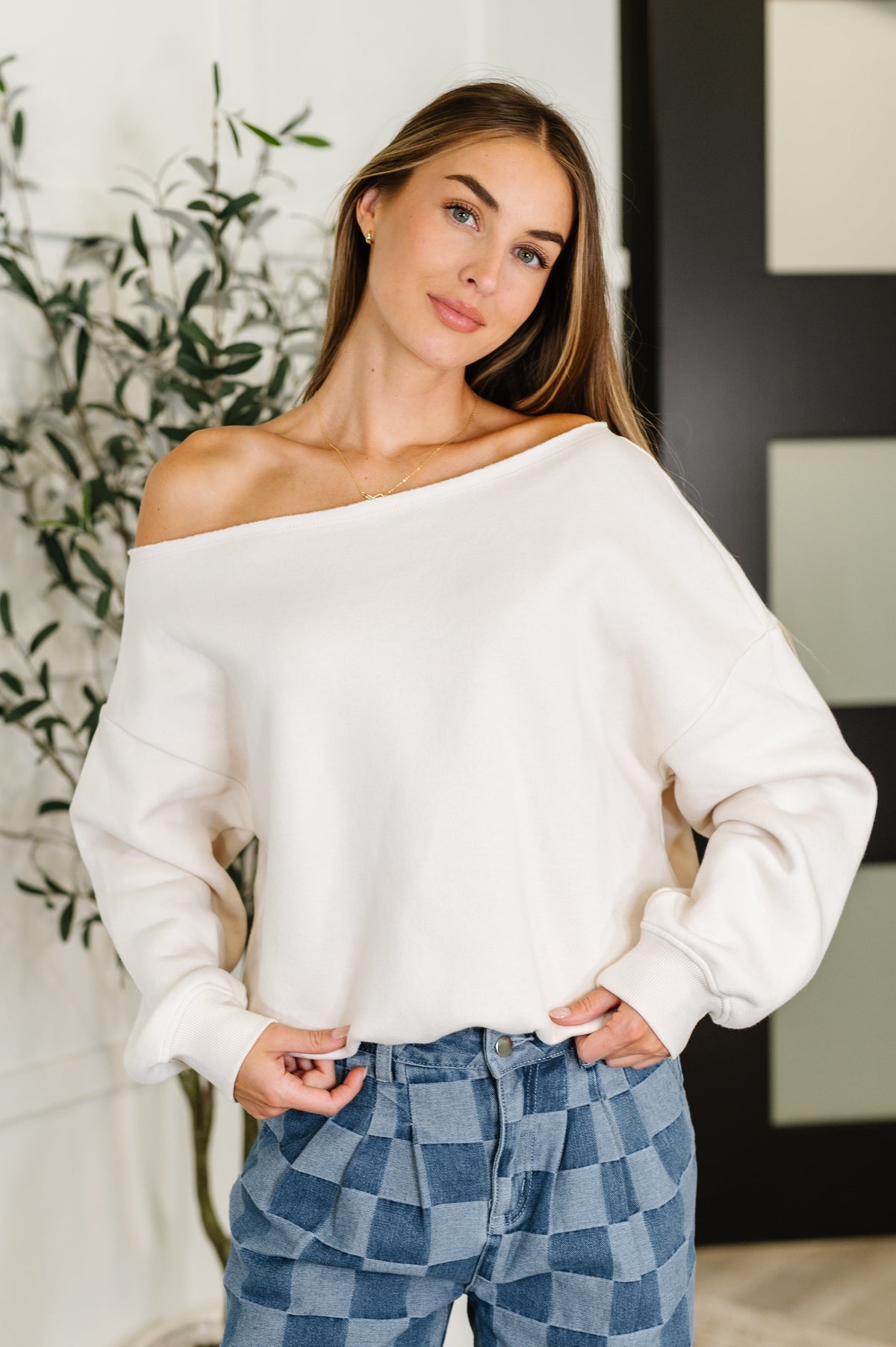 Woman wearing a white off-shoulder sweater and blue checkered pants indoors.