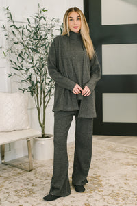 Woman wearing a gray outfit standing in a room with a plant and decorative wall.