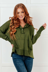 A woman with long wavy red hair wearing an olive green long-sleeve polo-style top and blue jeans, standing against a white paneled wall.