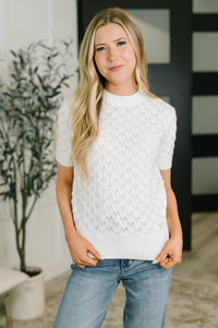 A woman with long blonde hair wearing a white short-sleeve knit sweater with a textured openwork pattern and light blue jeans, standing in a modern indoor setting.