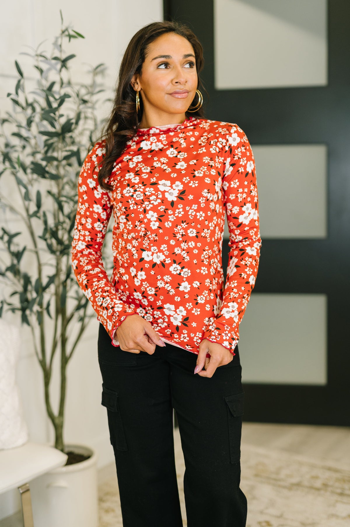 Woman wearing a red floral blouse standing indoors with a plant and wall art in the background
