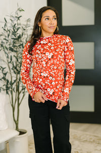 Woman wearing a red floral blouse standing indoors with a plant and wall art in the background