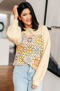 Woman wearing a beige sweater with a colorful granny square pattern over a plain top.
